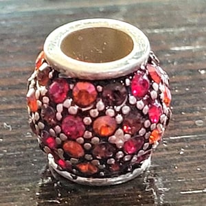 May include: A small, decorative bead with a silver-colored rim and a hollow center. The bead is covered in a mosaic of red, orange, and purple gemstones. The bead is likely used for jewelry making or other crafts.