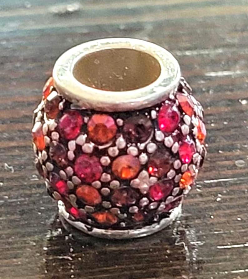 May include: A small, decorative bead with a silver-colored rim and a hollow center. The bead is covered in a mosaic of red, orange, and purple gemstones. The bead is likely used for jewelry making or other crafts.