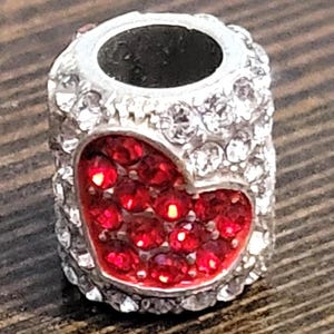 May include: A silver-colored charm bead with a heart-shaped design filled with red rhinestones. The bead is encrusted with clear rhinestones around the edges and has a dark hole through the center.