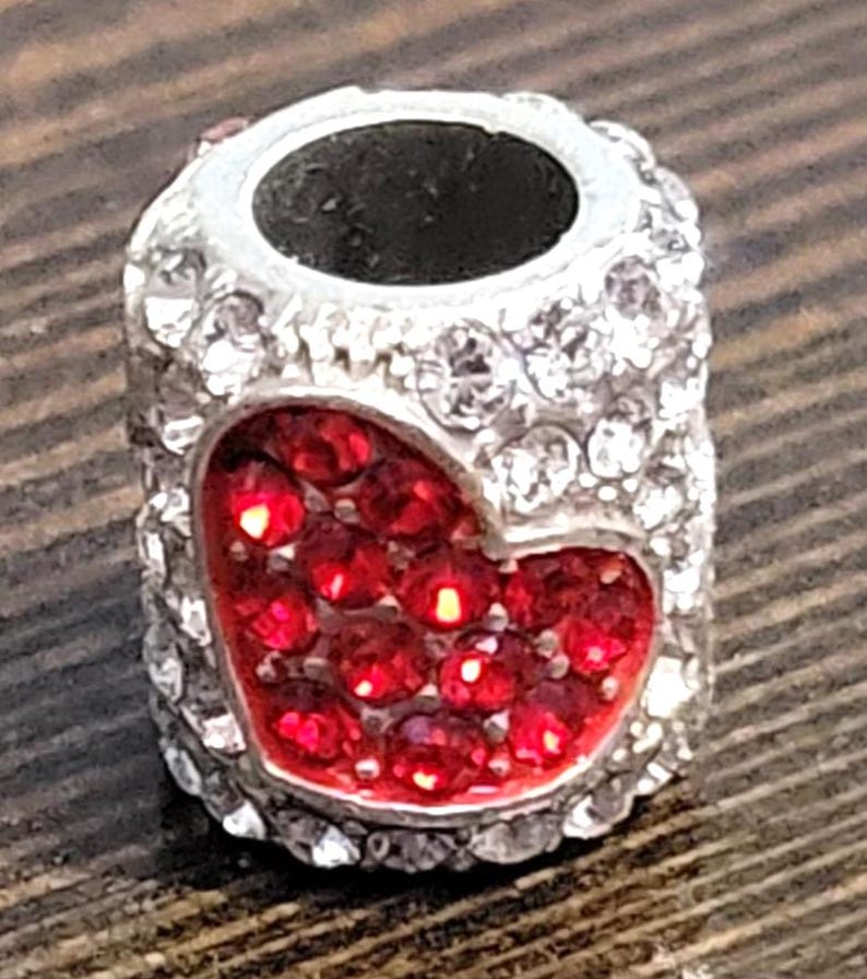 May include: A silver-colored charm bead with a heart-shaped design filled with red rhinestones. The bead is encrusted with clear rhinestones around the edges and has a dark hole through the center.