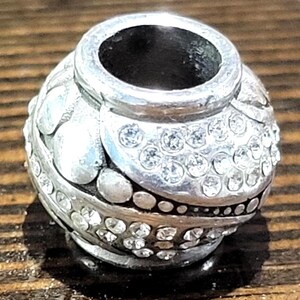 May include: A silver-toned bead with a rounded shape, adorned with clear, sparkling crystals. The bead features a textured surface with raised floral and dot patterns. The top opening is a smooth, unadorned silver. A decorative jewelry component.