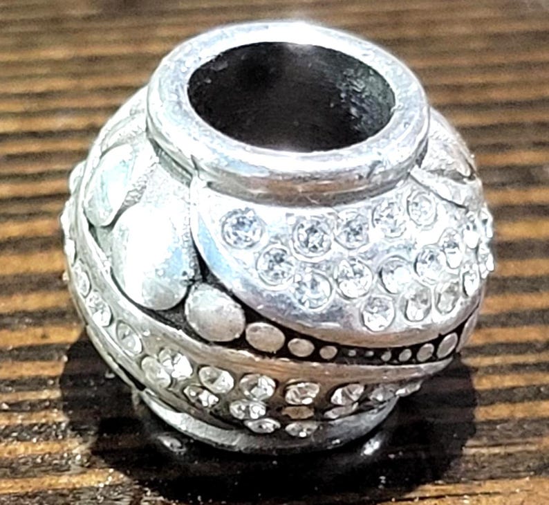 May include: A silver-toned bead with a rounded shape, adorned with clear, sparkling crystals. The bead features a textured surface with raised floral and dot patterns. The top opening is a smooth, unadorned silver. A decorative jewelry component.