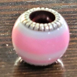 May include: A pink and white striped bead with a silver-colored, textured top. The bead has a dark red interior and is likely for jewelry making. The bead is oval-shaped and sits on a wooden surface.