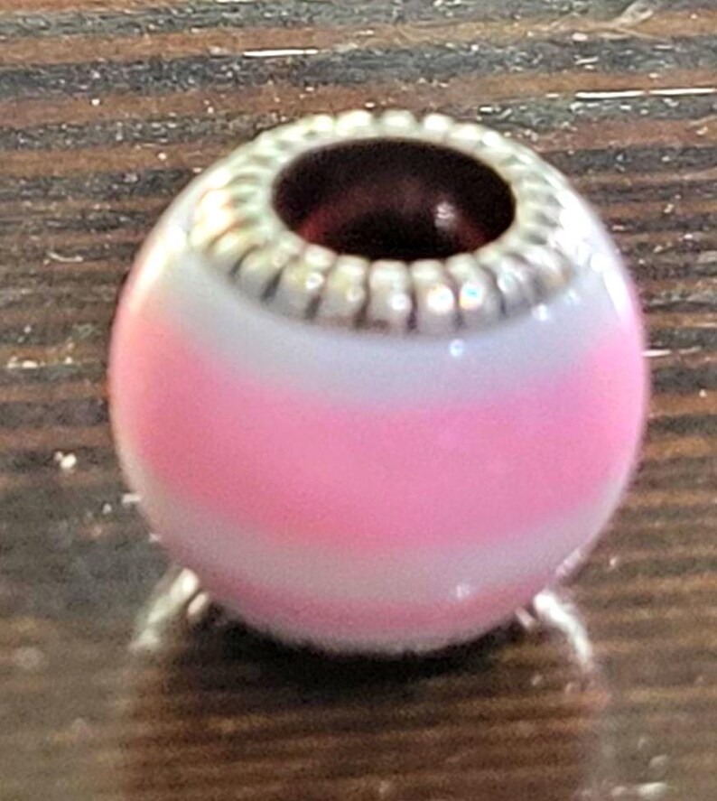 May include: A pink and white striped bead with a silver-colored, textured top. The bead has a dark red interior and is likely for jewelry making. The bead is oval-shaped and sits on a wooden surface.