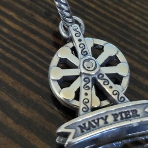 Puede incluir: Un charm plateado con un dise&ntilde;o de noria. El charm tiene una pancarta que dice "NAVY PIER". La noria tiene un dise&ntilde;o detallado con elementos decorativos. El charm est&aacute; suspendido de un lazo retorcido.