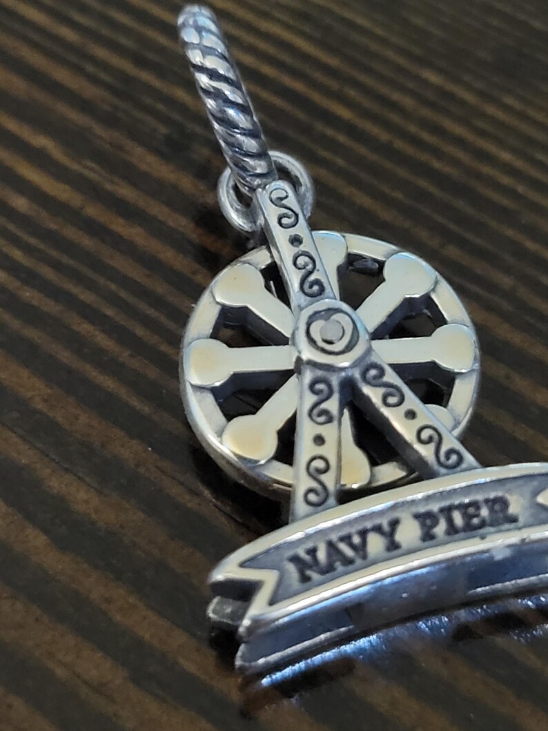 Puede incluir: Un charm plateado con un dise&ntilde;o de noria. El charm tiene una pancarta que dice "NAVY PIER". La noria tiene un dise&ntilde;o detallado con elementos decorativos. El charm est&aacute; suspendido de un lazo retorcido.