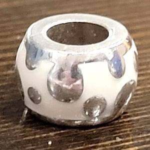 May include: A silver and white bead with a large central hole. The bead has a white base with silver-colored, irregular, droplet-shaped accents. The bead is likely made of metal and enamel, and is designed to be strung on a bracelet or necklace.