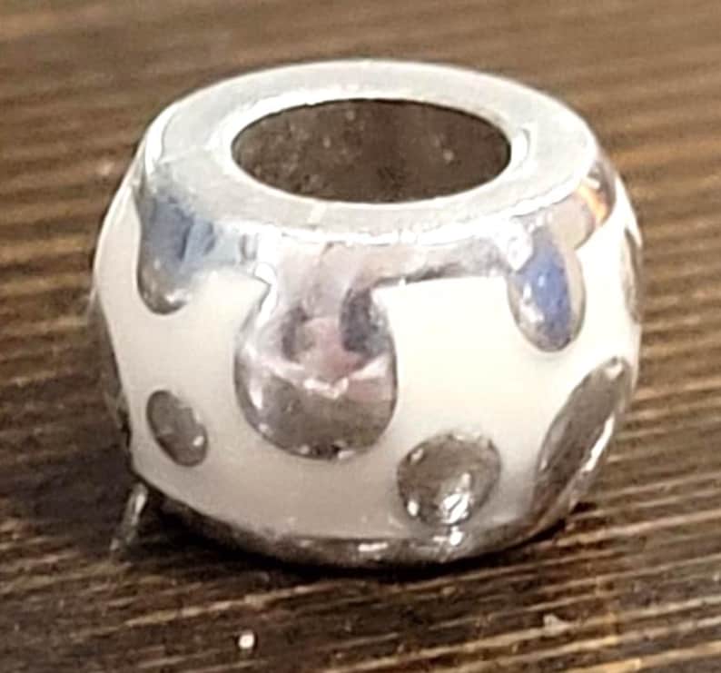 May include: A silver and white bead with a large central hole. The bead has a white base with silver-colored, irregular, droplet-shaped accents. The bead is likely made of metal and enamel, and is designed to be strung on a bracelet or necklace.