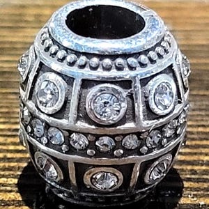 May include: Silver-toned bead with a geometric design, featuring clear, sparkling crystals. The bead has a cylindrical shape with a large central hole and is decorated with multiple rows of crystals and geometric patterns. The bead is likely used for jewelry making.