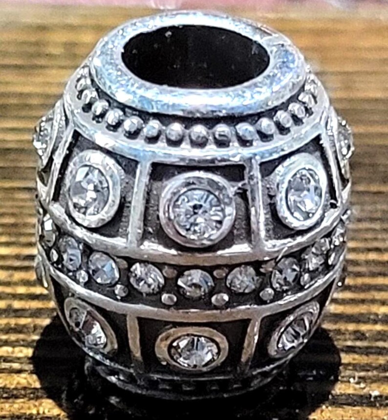 May include: Silver-toned bead with a geometric design, featuring clear, sparkling crystals. The bead has a cylindrical shape with a large central hole and is decorated with multiple rows of crystals and geometric patterns. The bead is likely used for jewelry making.