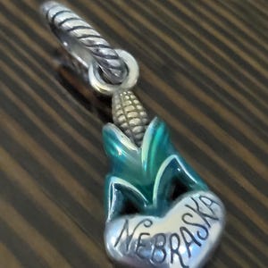 Puede incluir: Un charm de plata con un dise&ntilde;o de mazorca de ma&iacute;z con detalles en verde y azul. El charm incluye un elemento en forma de coraz&oacute;n con la palabra "NEBRASKA" grabada. Tiene una argolla retorcida de plata.