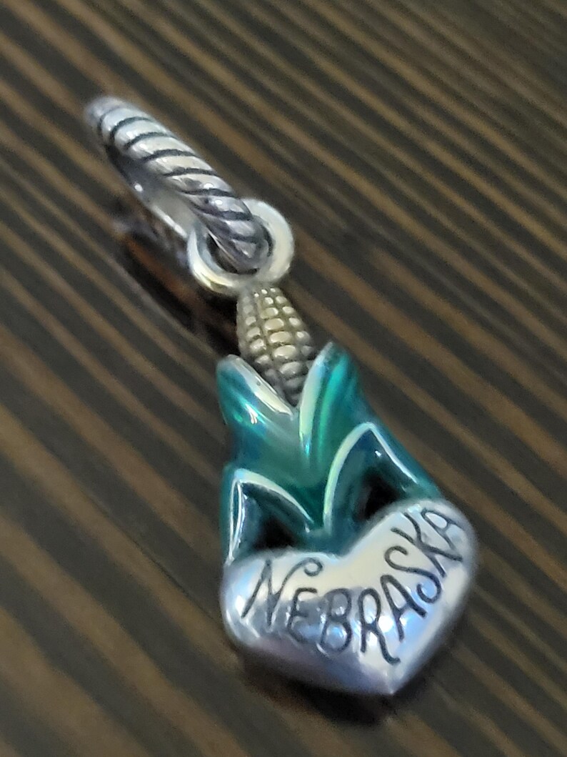 Puede incluir: Un charm de plata con un dise&ntilde;o de mazorca de ma&iacute;z con detalles en verde y azul. El charm incluye un elemento en forma de coraz&oacute;n con la palabra "NEBRASKA" grabada. Tiene una argolla retorcida de plata.