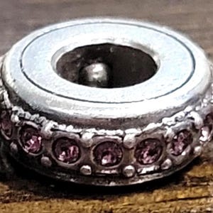 May include: A silver-toned charm with a circular design. The charm features a ring of small, pink gemstones around the base. The center has a hole, and the charm has a vintage or antique appearance.