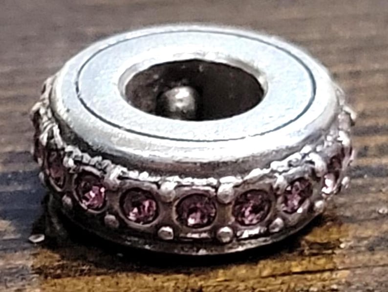 May include: A silver-toned charm with a circular design. The charm features a ring of small, pink gemstones around the base. The center has a hole, and the charm has a vintage or antique appearance.