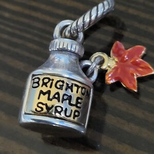 Puede incluir: Un dije plateado en forma de botella de jarabe de arce con las palabras "BRIGHTON MAPLE SYRUP" en letras negras. Un dije de hoja de arce roja y dorada cuelga de la botella. El dije tiene un lazo plateado retorcido.