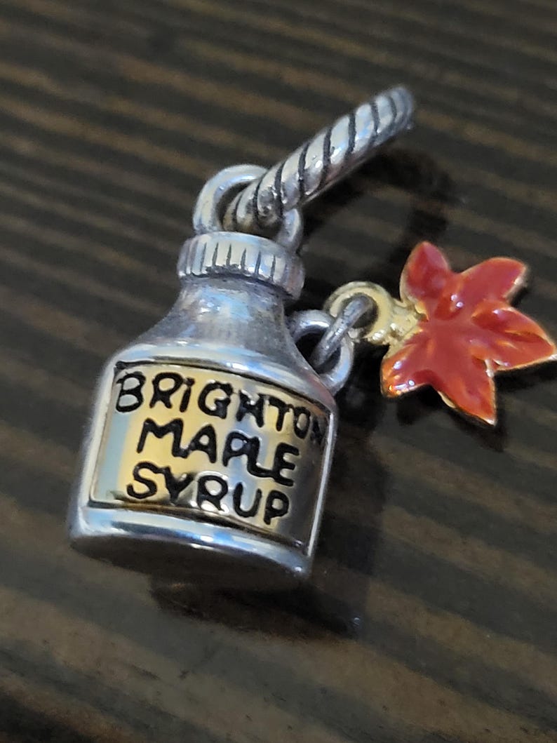 Puede incluir: Un dije plateado en forma de botella de jarabe de arce con las palabras "BRIGHTON MAPLE SYRUP" en letras negras. Un dije de hoja de arce roja y dorada cuelga de la botella. El dije tiene un lazo plateado retorcido.