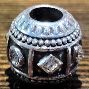 May include: Silver-toned bead with a dark, textured base. The bead features a decorative pattern with clear, sparkling accents. The top has a large opening, and the design includes geometric shapes and beaded details.