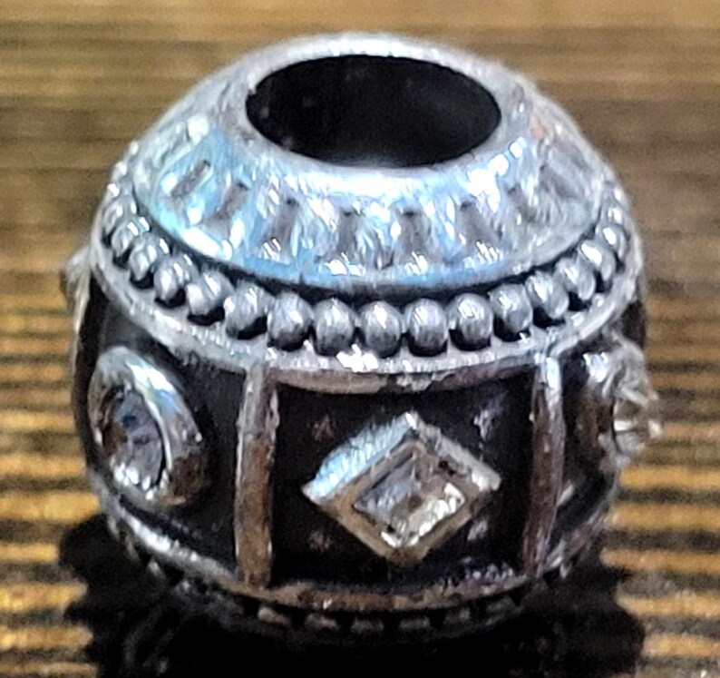 May include: Silver-toned bead with a dark, textured base. The bead features a decorative pattern with clear, sparkling accents. The top has a large opening, and the design includes geometric shapes and beaded details.