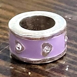 May include: A silver charm with a lavender enamel band. The band features three clear, heart-shaped accents. The charm has a silver rim and a dark interior. The charm is designed to be a bead for a bracelet or necklace.
