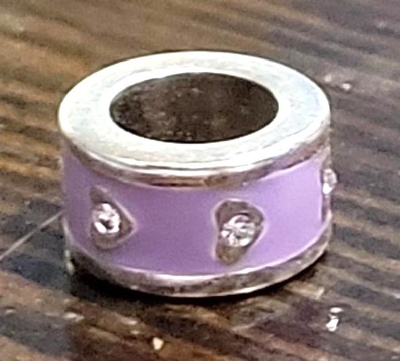 May include: A silver charm with a lavender enamel band. The band features three clear, heart-shaped accents. The charm has a silver rim and a dark interior. The charm is designed to be a bead for a bracelet or necklace.