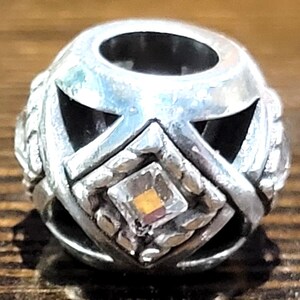 May include: A silver-toned bead with a geometric design. The bead features cut-out shapes and a central square with a small, multi-colored gem. The bead has a hole running through the center.