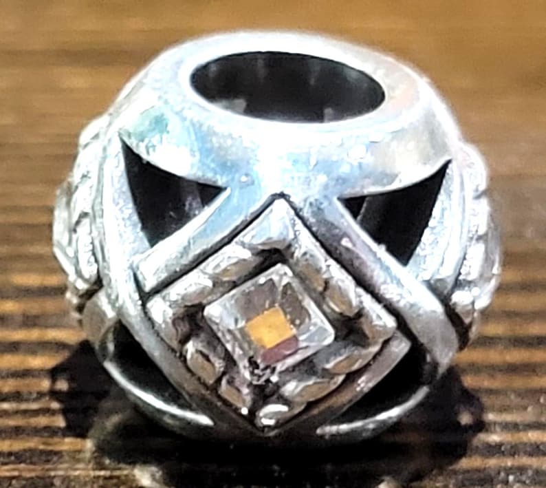 May include: A silver-toned bead with a geometric design. The bead features cut-out shapes and a central square with a small, multi-colored gem. The bead has a hole running through the center.