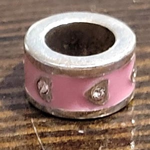 May include: A silver-toned charm with a pink enamel band. The charm features three clear, sparkling accents. The charm has a circular design with a hollow center, suitable for a bracelet or necklace.