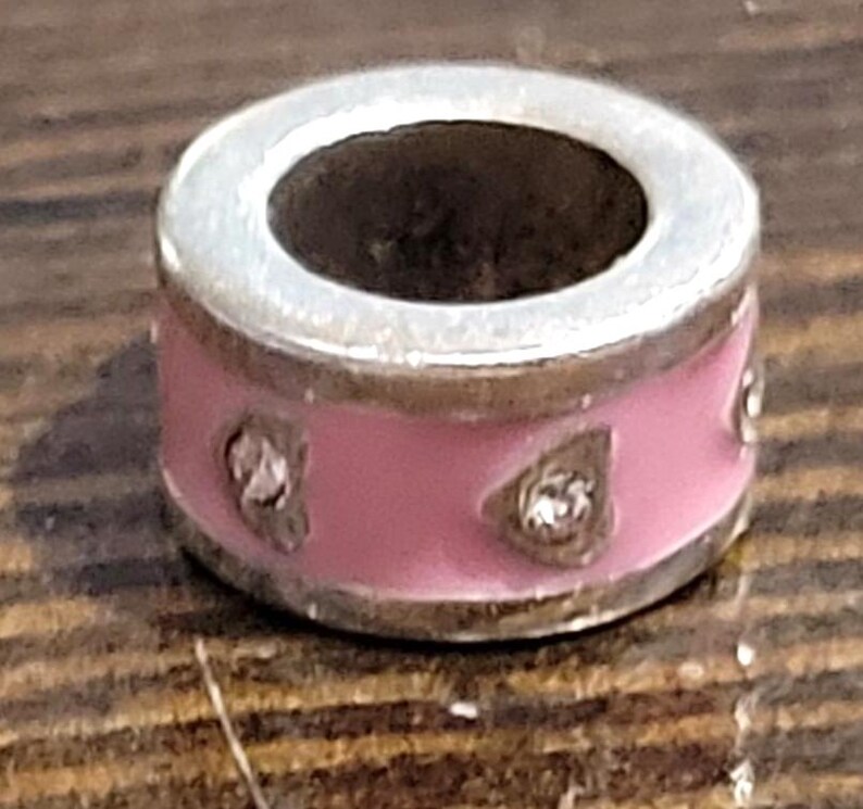May include: A silver-toned charm with a pink enamel band. The charm features three clear, sparkling accents. The charm has a circular design with a hollow center, suitable for a bracelet or necklace.