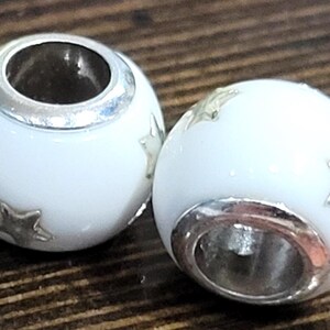 May include: Two white, spherical beads with silver-colored accents and star-shaped embellishments. The beads have a hole running through the center and are set against a dark wood grain background. These beads are likely for jewelry making.