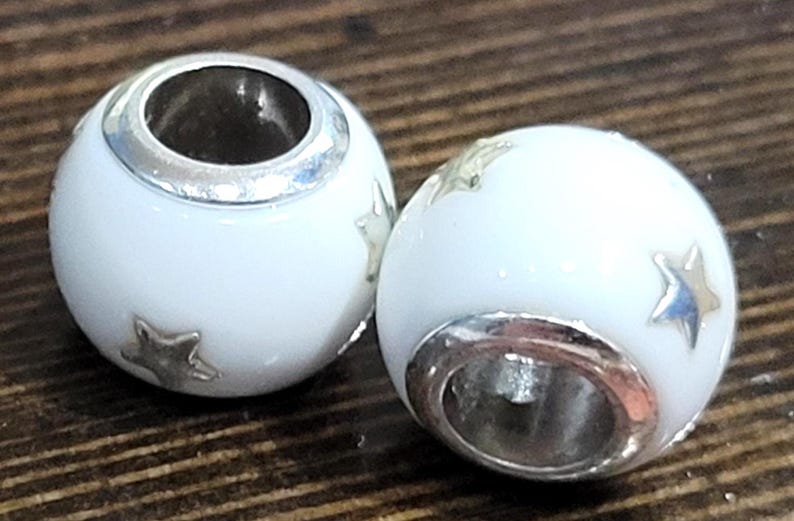 May include: Two white, spherical beads with silver-colored accents and star-shaped embellishments. The beads have a hole running through the center and are set against a dark wood grain background. These beads are likely for jewelry making.