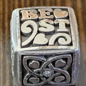 May include: A silver-toned charm with the word "BEST" in gold lettering and heart accents. The bottom section features an intricate Celtic knot design. The charm is likely for a bracelet or necklace.