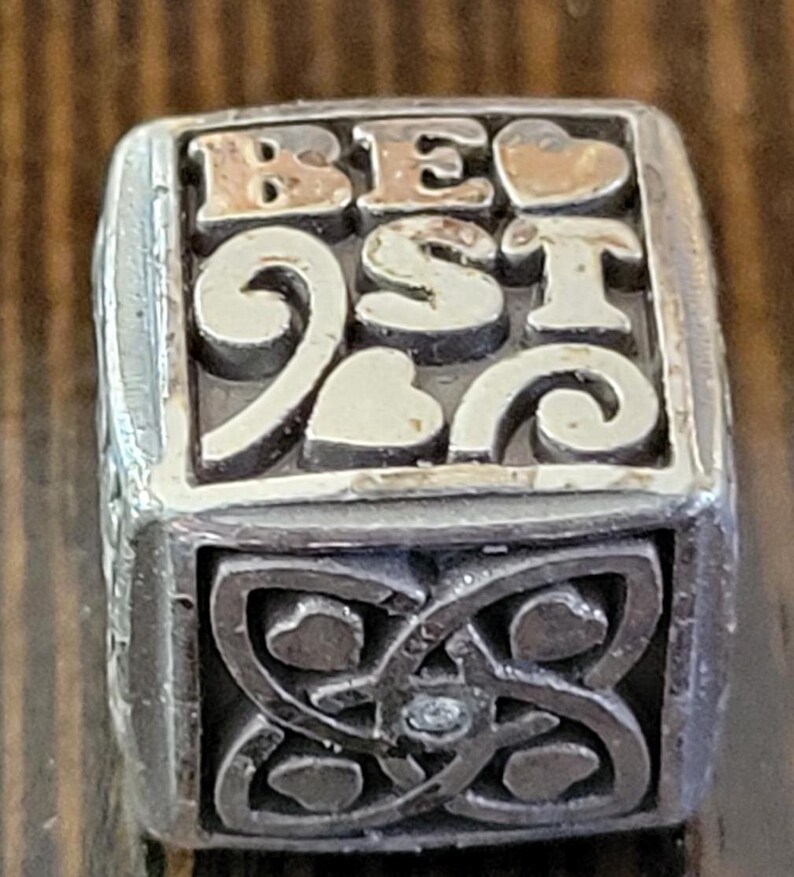 May include: A silver-toned charm with the word "BEST" in gold lettering and heart accents. The bottom section features an intricate Celtic knot design. The charm is likely for a bracelet or necklace.