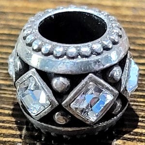 May include: A silver-toned bead with clear, square-cut crystals and small round accents. The bead has a dark interior and a decorative, textured design. This jewelry component is suitable for bracelets and necklaces.