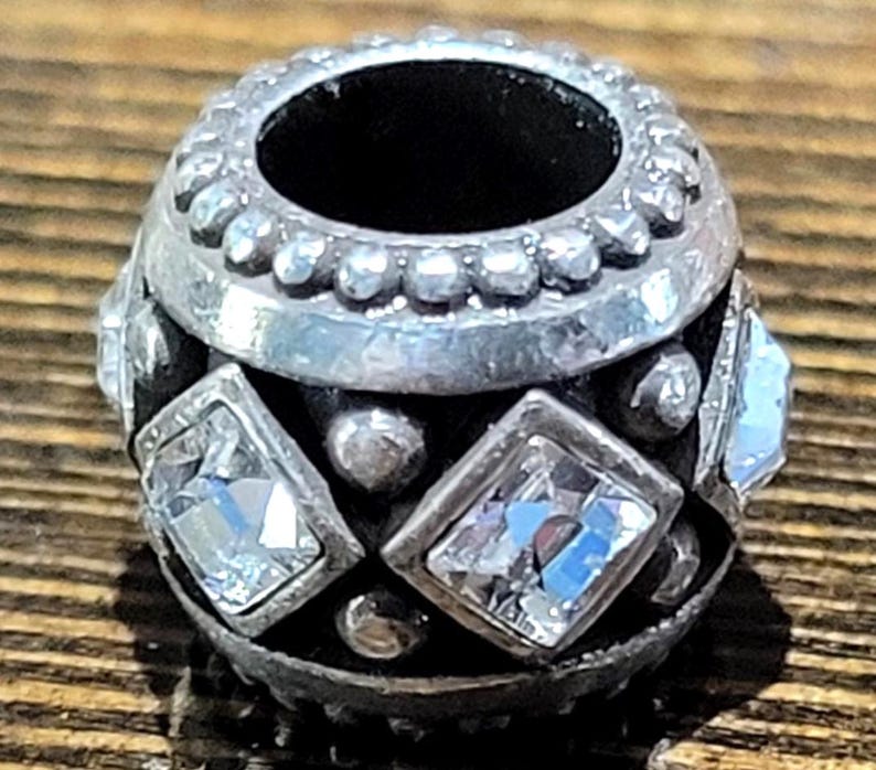 May include: A silver-toned bead with clear, square-cut crystals and small round accents. The bead has a dark interior and a decorative, textured design. This jewelry component is suitable for bracelets and necklaces.