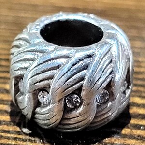 May include: Silver-toned bead with a woven design and three clear, round crystals. The bead has a large central hole and is likely for jewelry making or charm bracelets. The bead is on a wooden surface.