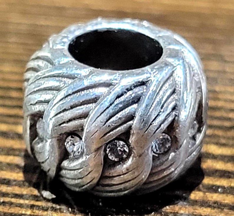 May include: Silver-toned bead with a woven design and three clear, round crystals. The bead has a large central hole and is likely for jewelry making or charm bracelets. The bead is on a wooden surface.