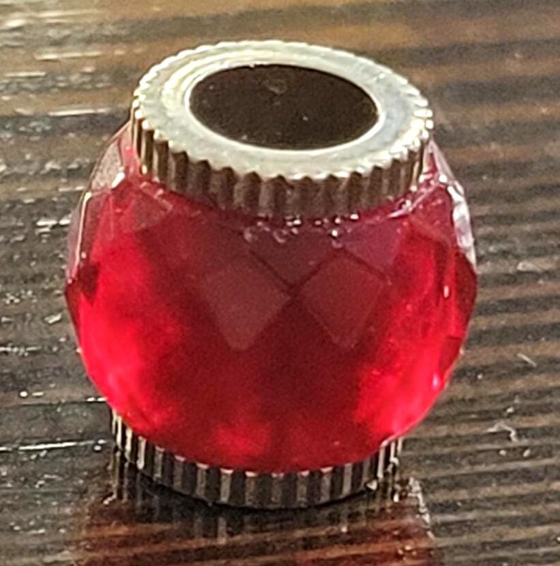 May include: A faceted, ruby-red bottle stopper with a silver-colored, ridged metal rim. The stopper has a cylindrical shape with a flat top and bottom. The glass is transparent, allowing light to pass through.