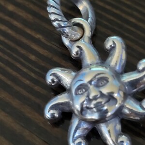 Puede incluir: Un dije plateado con un sol sonriente con rayos. El dije tiene un lazo texturizado para sujetarlo. La cara del sol tiene detalles, incluyendo ojos, nariz y boca sonriente. El dije es probablemente para un collar o pulsera.
