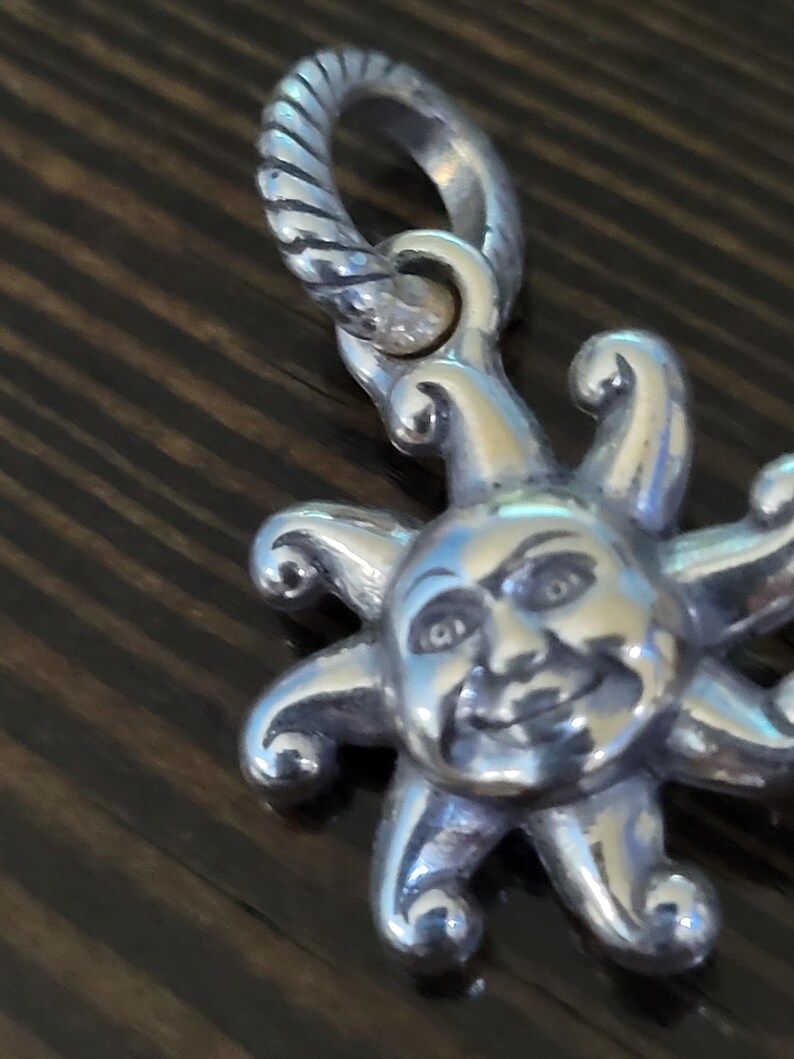 Puede incluir: Un dije plateado con un sol sonriente con rayos. El dije tiene un lazo texturizado para sujetarlo. La cara del sol tiene detalles, incluyendo ojos, nariz y boca sonriente. El dije es probablemente para un collar o pulsera.