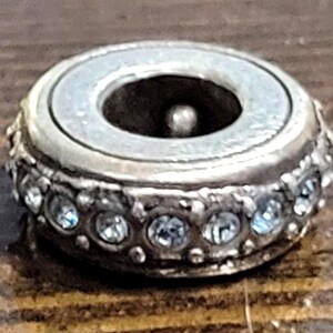 May include: A silver-toned bead with a central hole and a ring of clear, sparkling crystals. The bead has a light-colored, pearlescent center and sits on a wooden surface. The bead is likely for jewelry making.