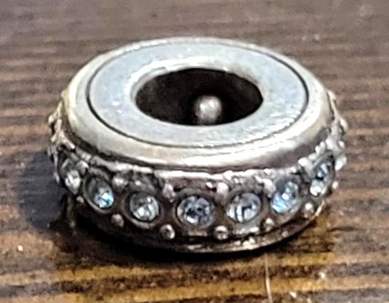 May include: A silver-toned bead with a central hole and a ring of clear, sparkling crystals. The bead has a light-colored, pearlescent center and sits on a wooden surface. The bead is likely for jewelry making.
