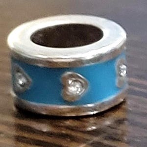 May include: A silver charm with a light blue band featuring three heart-shaped embellishments. The charm has a cylindrical shape with a dark interior and is designed for bracelets or necklaces.