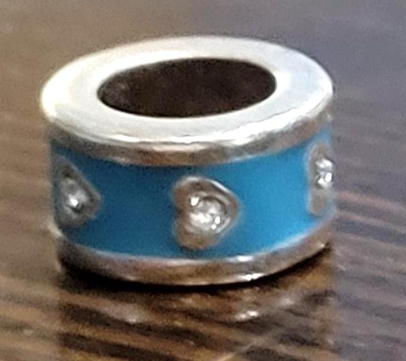 May include: A silver charm with a light blue band featuring three heart-shaped embellishments. The charm has a cylindrical shape with a dark interior and is designed for bracelets or necklaces.