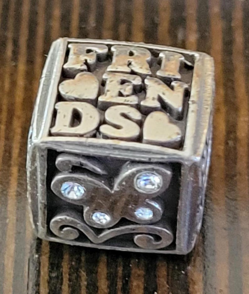 May include: A silver-toned, rectangular charm with the word "FRIENDS" in raised letters. The charm features heart accents and a butterfly design with clear crystal embellishments, perfect for a bracelet or necklace.