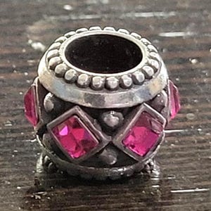 May include: A silver-toned bead with a central opening and a decorative design. The bead features alternating pink, square-cut gemstones and small, round embellishments. The top and bottom edges have a beaded border.