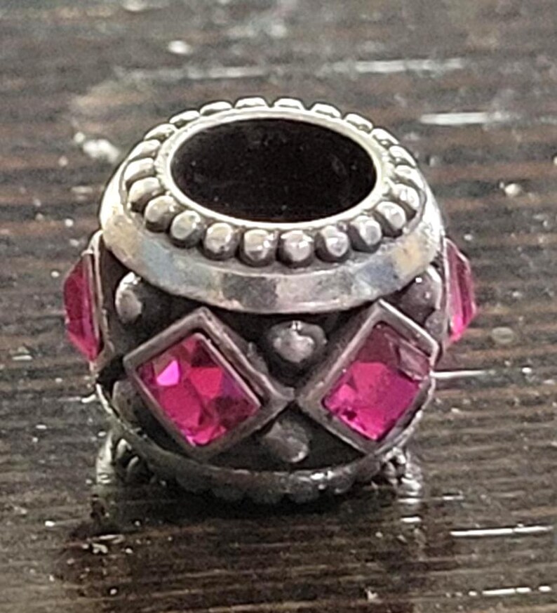 May include: A silver-toned bead with a central opening and a decorative design. The bead features alternating pink, square-cut gemstones and small, round embellishments. The top and bottom edges have a beaded border.