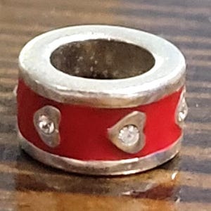 May include: A silver-toned charm with a red enamel band. The band is decorated with heart-shaped accents, each containing a small, clear gemstone. The charm has a central opening and is designed to be used with a bracelet or necklace.