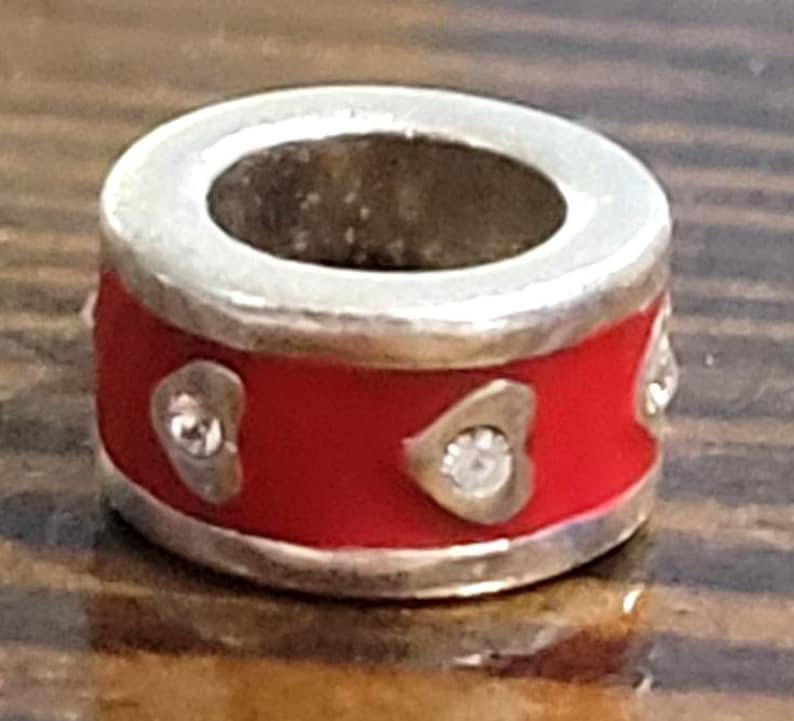 May include: A silver-toned charm with a red enamel band. The band is decorated with heart-shaped accents, each containing a small, clear gemstone. The charm has a central opening and is designed to be used with a bracelet or necklace.