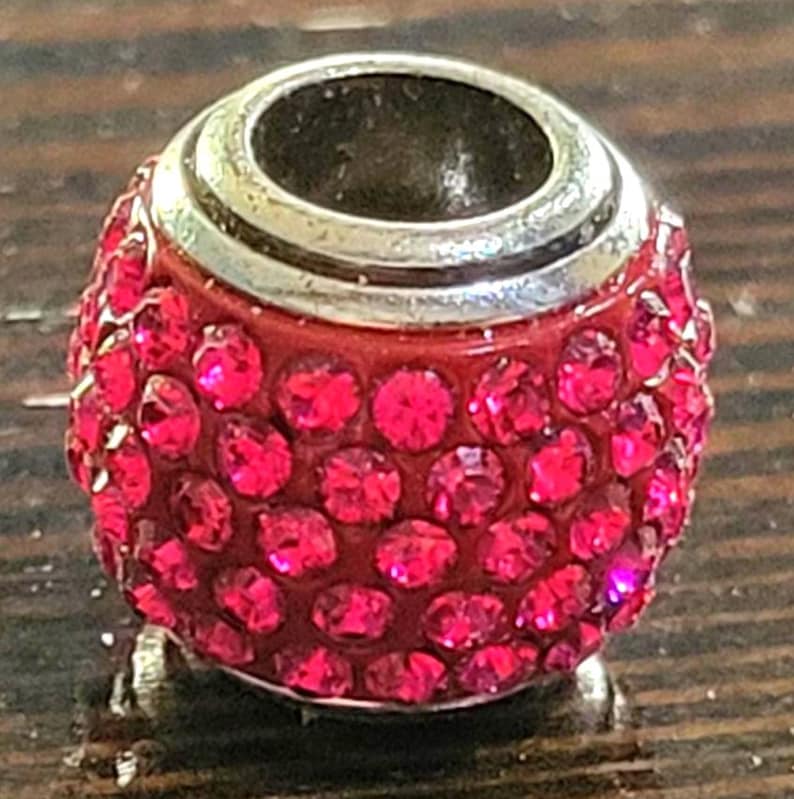 May include: A red charm bead with a silver-colored top and a base. The bead is covered in small, sparkling, red crystals. The bead is round and has a hole through the center.