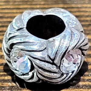 May include: A silver-toned bead with a central hole and swirling, textured design. Two clear, faceted gemstones are set into the bead, one on each side. The bead has a vintage or antique appearance.